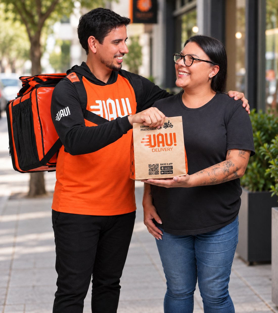 Na calçada de uma rua comercial, entregador do Uau! Delivery, de uniforme laranja e bag térmica nas costas, entrega uma sacola da marca para uma cliente sorridente